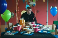 un homme habillé en noir assis à une table sur laquelle il y a des assiettes en plastique bleu et des verres roses. La table est entourée de ballon.